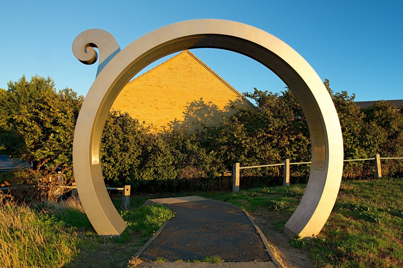 A tall steel ring sculpture over a path, framing a sunlit gable behind hedges