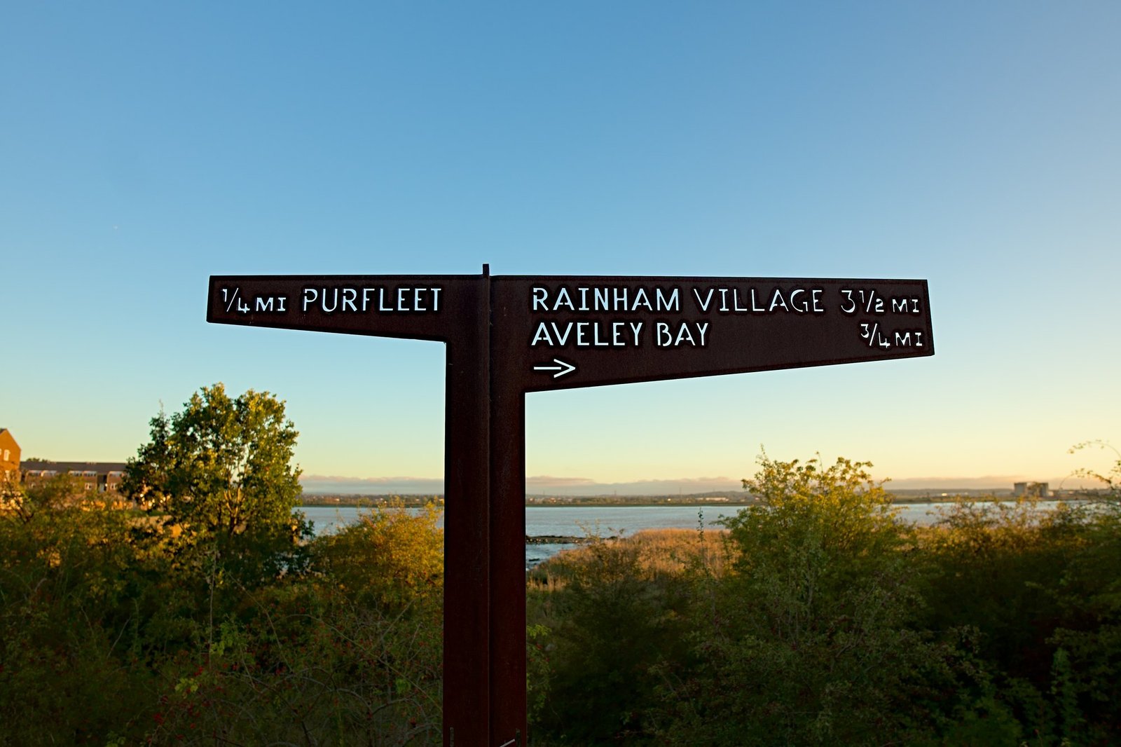 A wooden fingerpost pointing to Purfleet, Rainham Village and Aveley Bay against a dusk sky